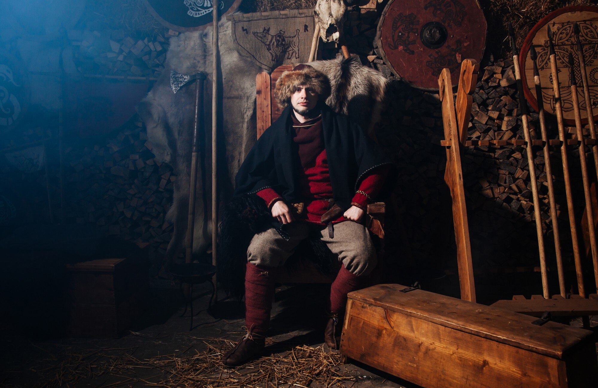 A man in a fur hat and dark cloak sits in a dim rustic interior with shields and wooden furnishings.