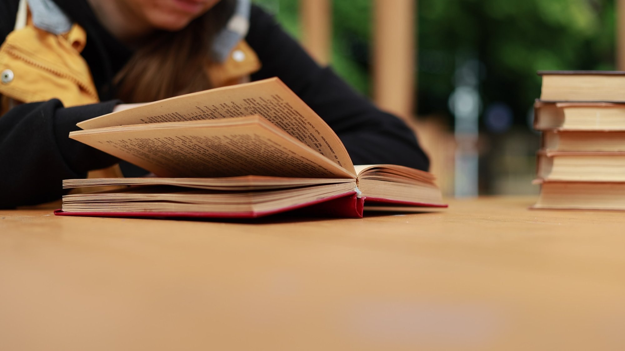 A woman reads an open book at a wooden desk beside a stack of books.