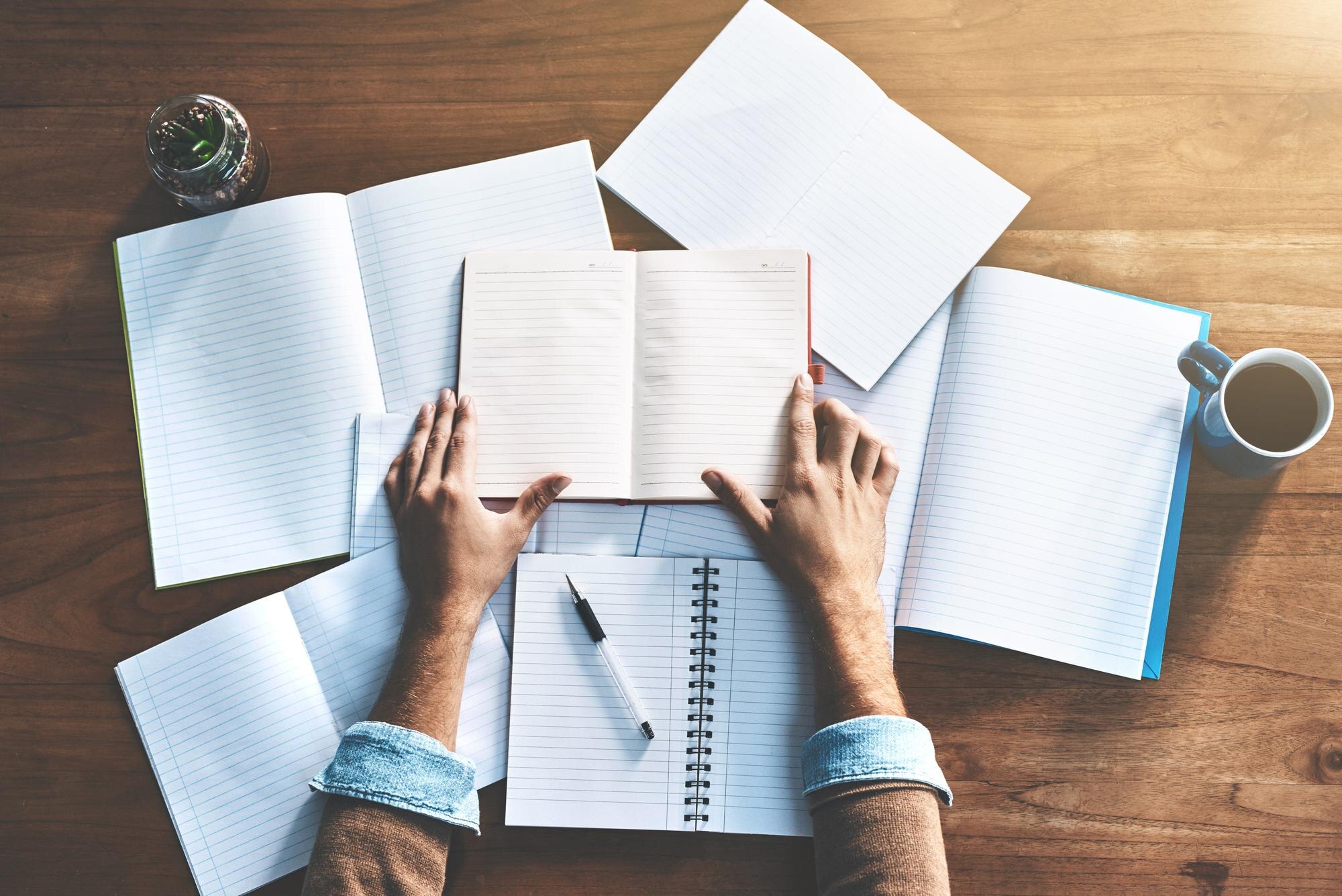 Overhead view of hands holding an open notebook among several notebooks on a wooden desk beside coffee.