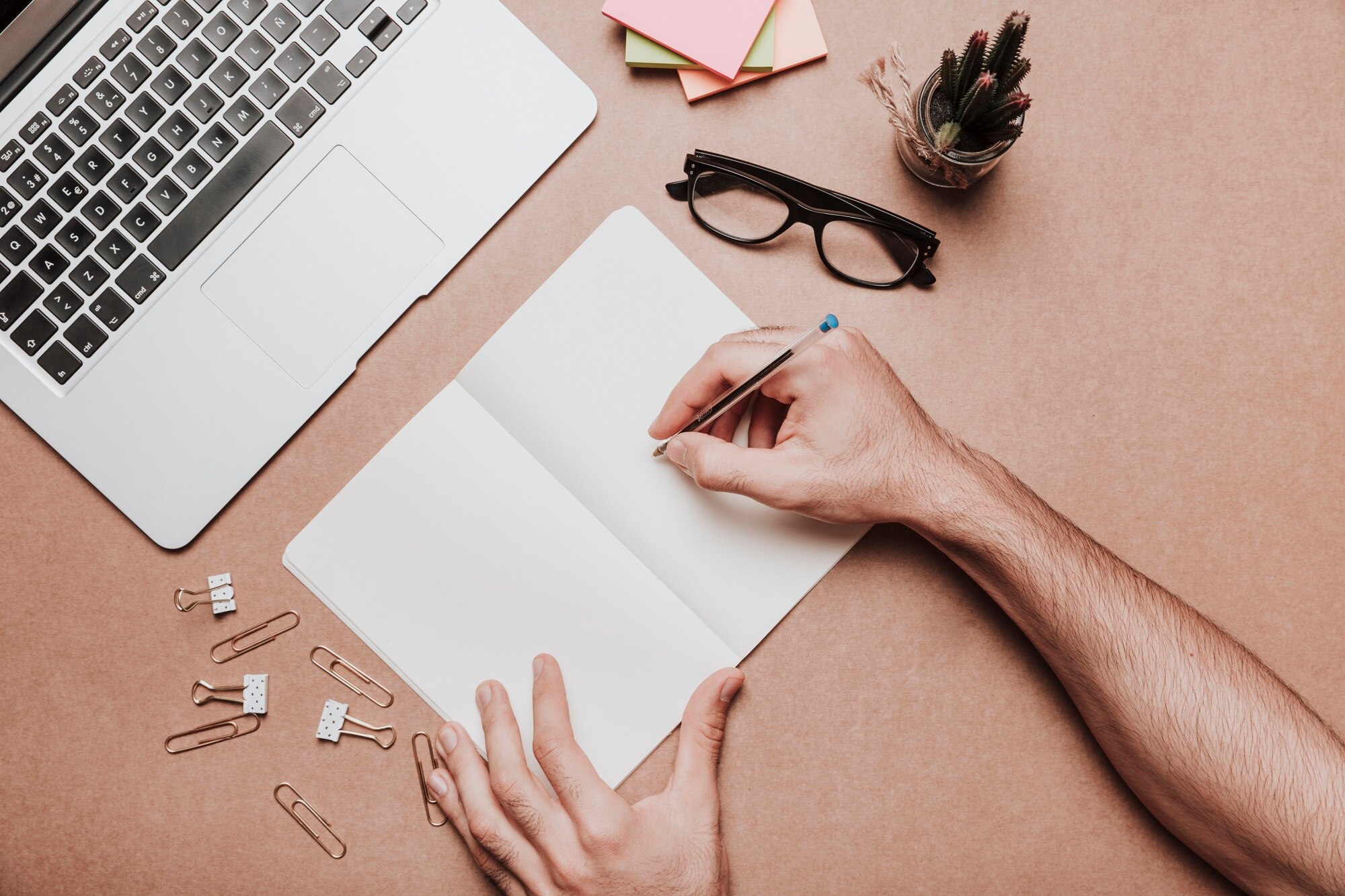 Hands holding a pencil over a blank notebook beside a laptop, pastel note cards, glasses and a small cactus.