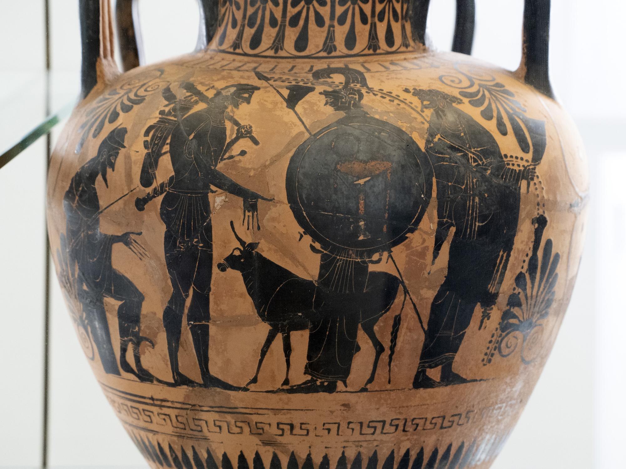 Black-figure pottery vessel decorated with warriors, a round shield and a dog behind glass.