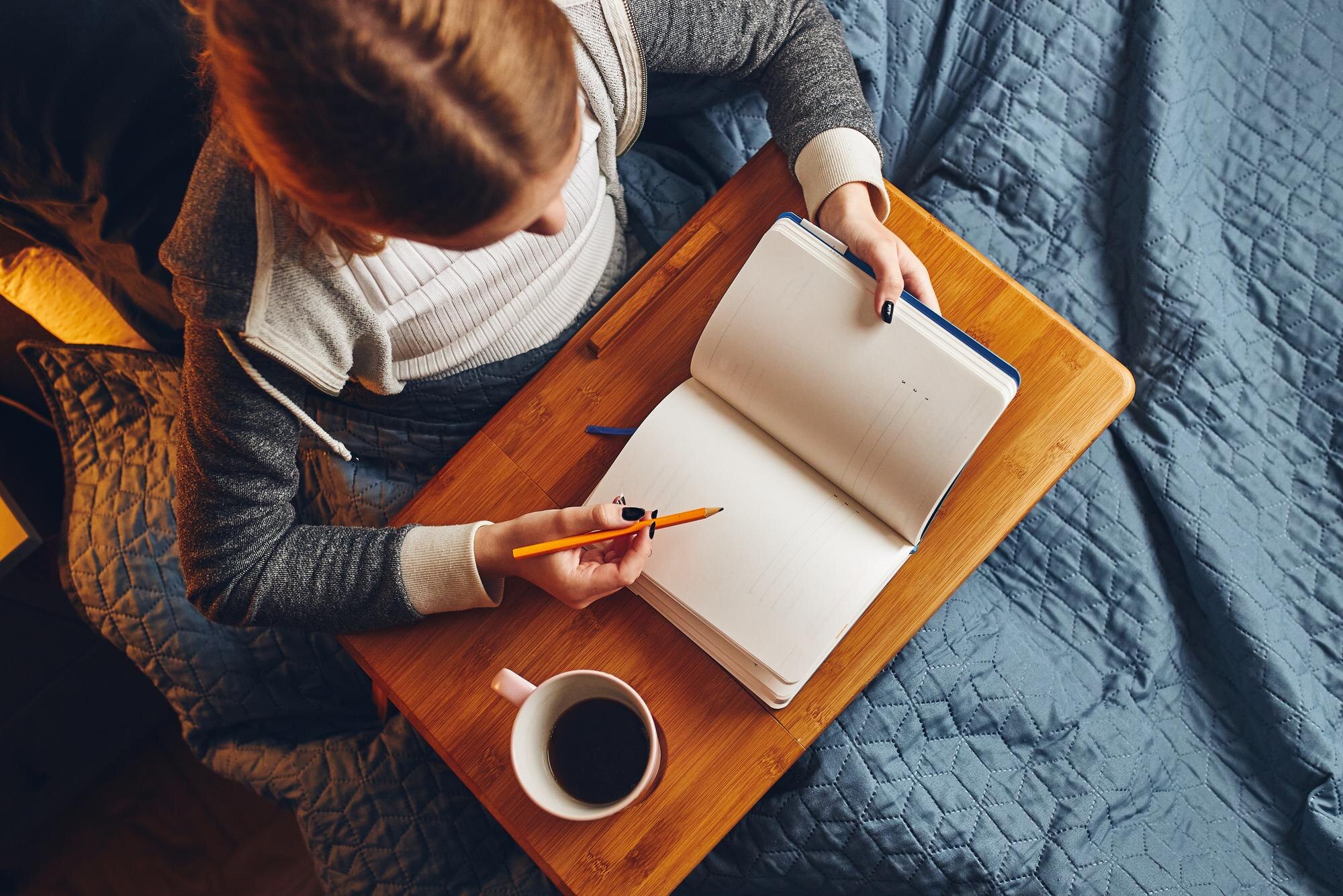 Woman holding a pencil over an open notebook on a wooden lap desk beside a cup of coffee.