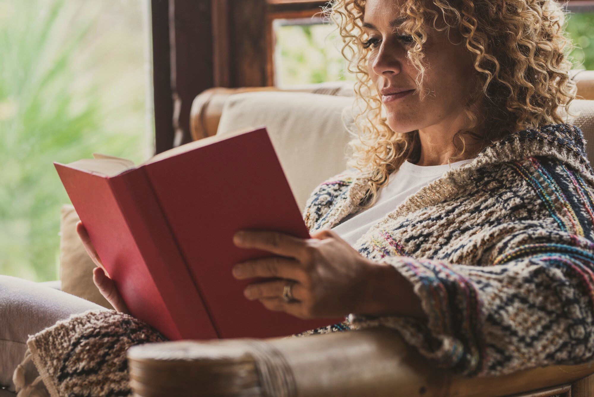 Woman sitting in a cushioned chair by a window, reading a book with a plain red cover.