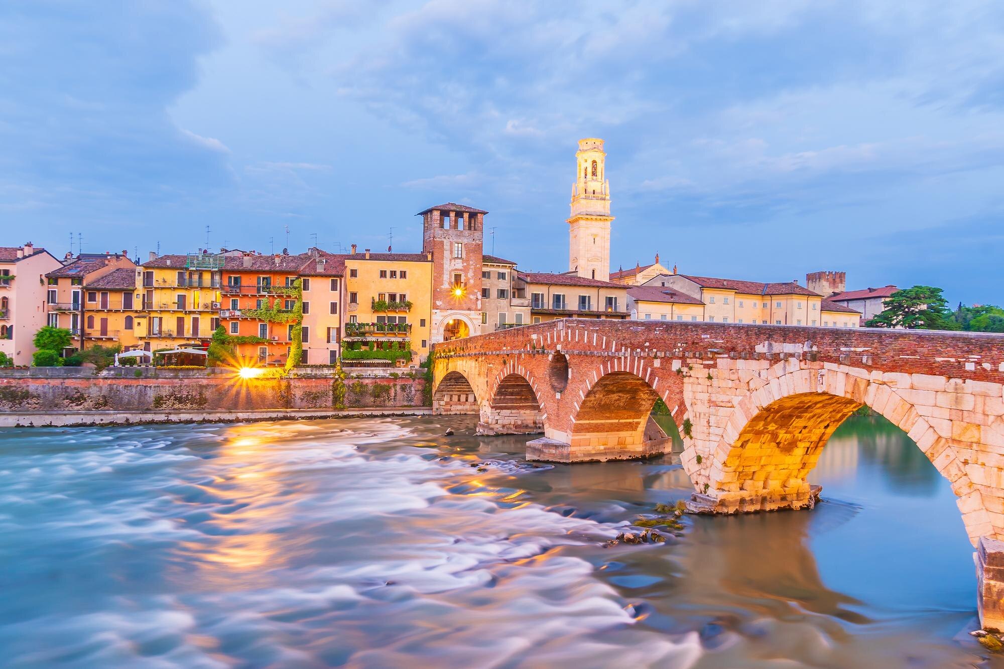 Stone arch bridge over the river beside colorful buildings and towers in Verona, Italy, at dusk.