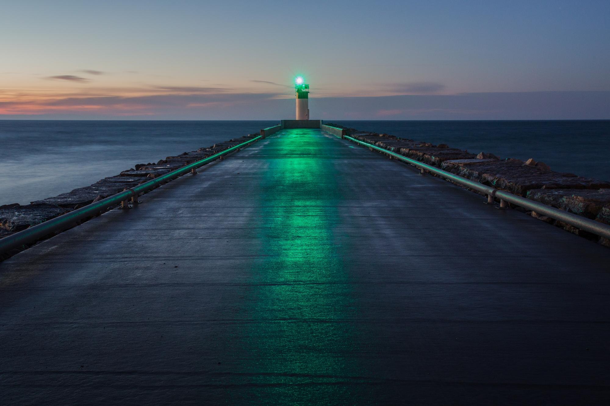 A green beacon glows at the end of a wet pier surrounded by dark water at dusk.