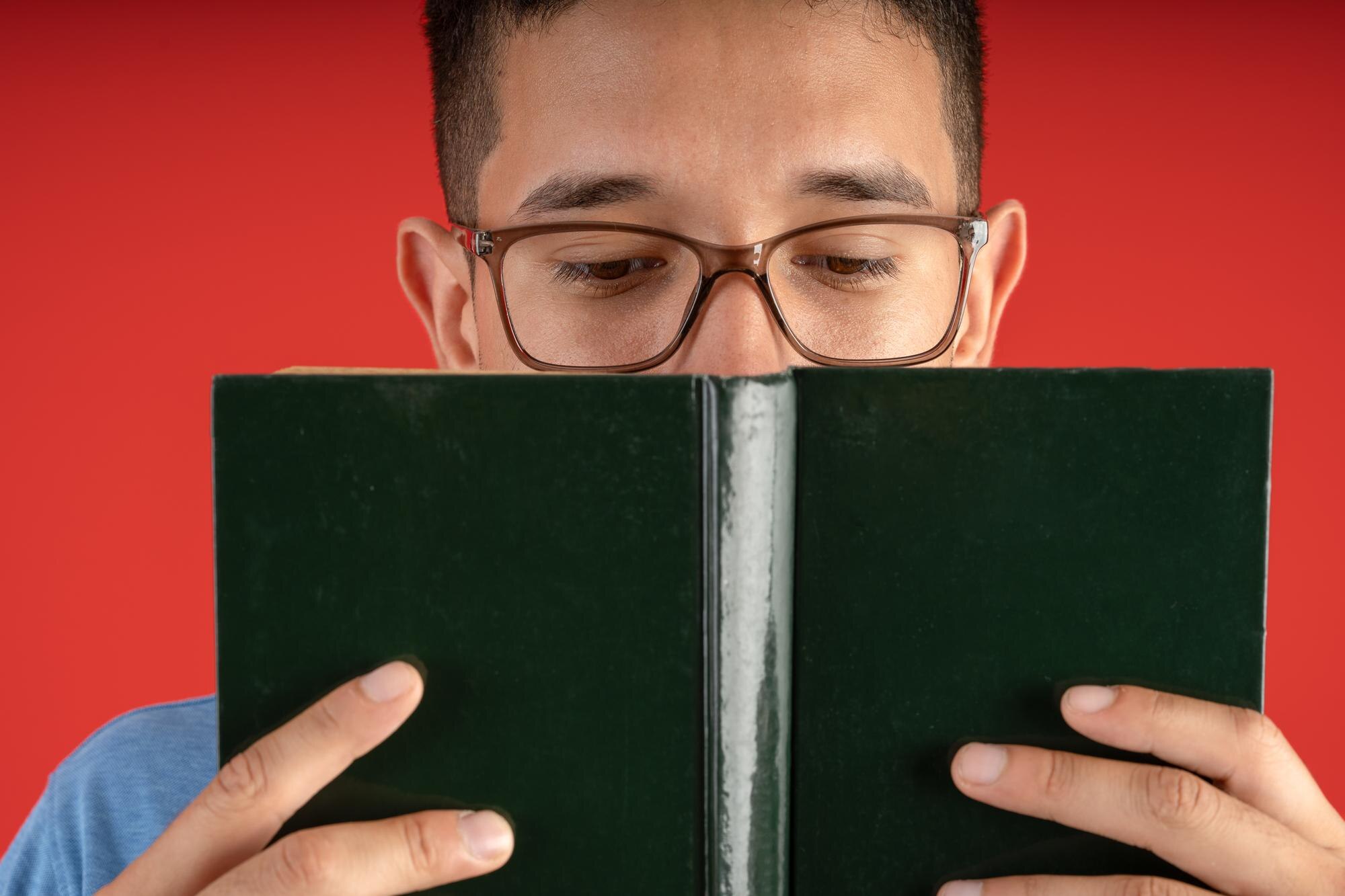 Man wearing glasses holds an open, unmarked dark green hardcover book in front of his face against a red background.